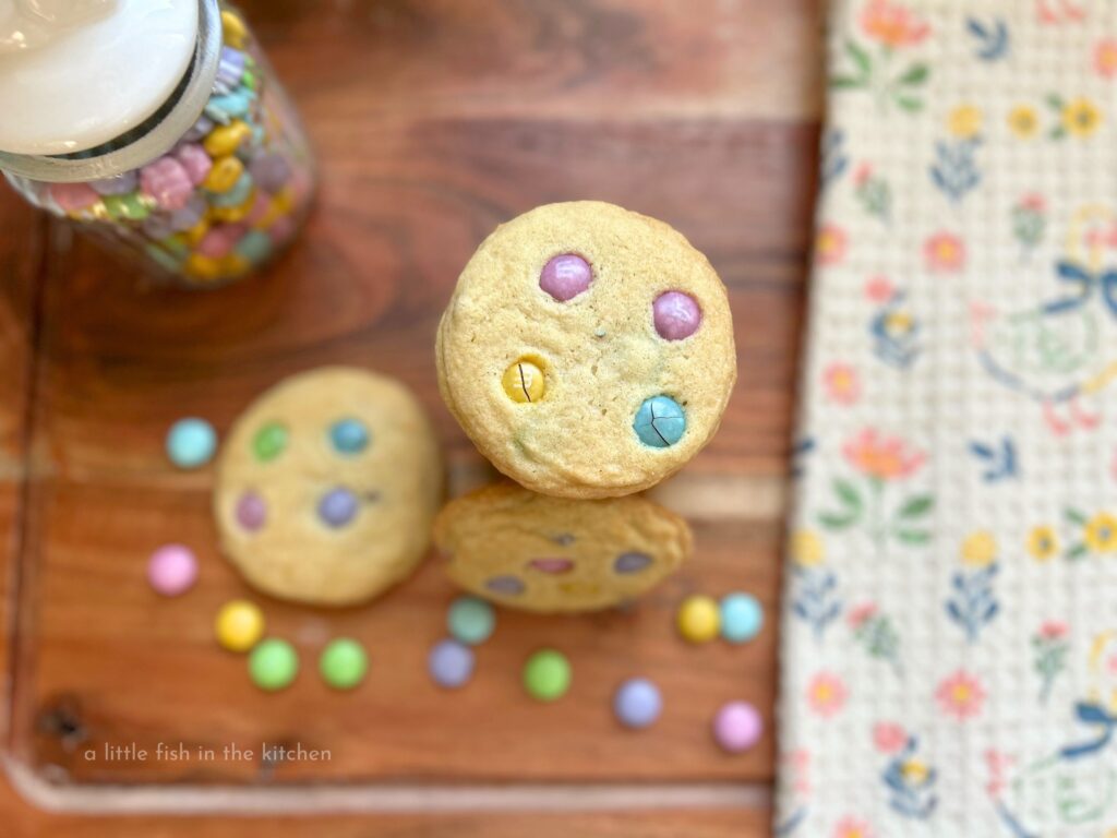 Buttery homemade cookies are studded with festive, colorful m&m candies. A few pastel colored chocolate candies are spread around a fresh-baked stack of golden cookies. A beige tea towel with a pasted floral pattern is next to the stack of cookies on a wodded cutting board and a small clear glass jar is filled with pastel m&m candies. 