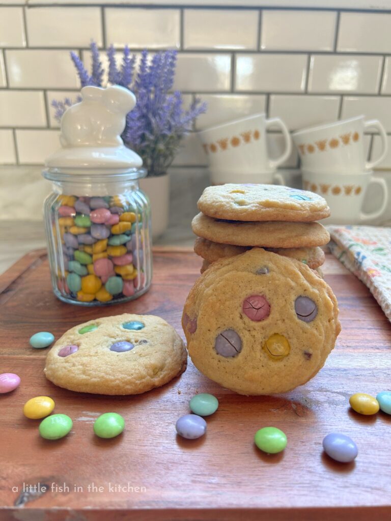 Buttery, homemade m&m cookies are stacked on a wooden cutting board. One cookie is laying flat and another cookie is on it's side leaning against the stack of eye-catching cookies. Several pastel-colored chocolate candies are sprinked around the cutting board. A small glass jar with a white ceramic topper shaped like a bunny is next to the fresh baked cookies, it's filled with colorful pastel m&m candies