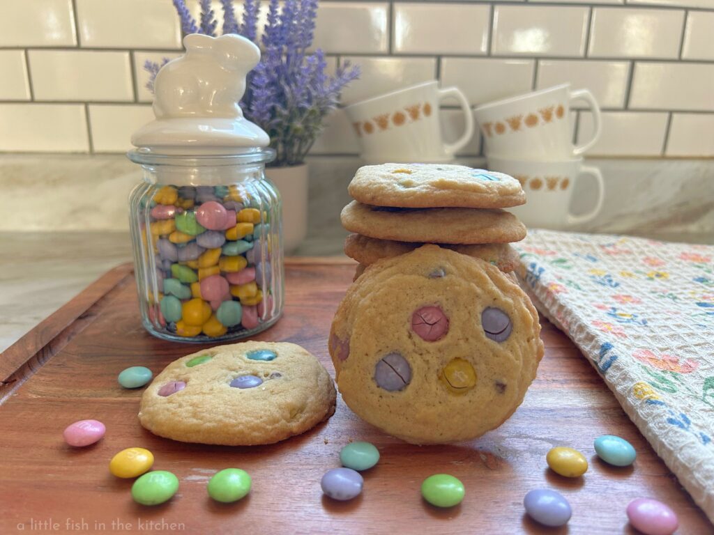 Buttery, homemade m&m cookies are stacked on a wooden cutting board. One cookie is laying flat and another cookie is on it's side leaning against the stack of eye-catching cookies. Several pastel-colored chocolate candies are sprinked around the cutting board. A small glass jar with a white ceramic topper shaped like a bunny is next to the fresh baked cookies, it's filled with colorful pastel m&m candies. Vintage Pyrex coffee cups with yellow sunflowers and a lilac plant are slightly blurred in the background for a decorative effect. 