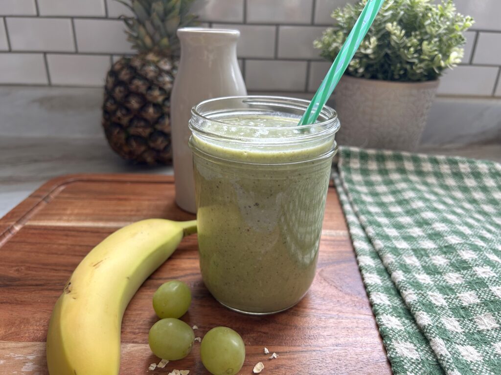 A single creamy green smoothie with oats is served in a clear glass mason jar. Bits of blended seeds are visible in the sage green-colored drink. The smoothie is served on a wooden cutting board with a whole yellow banana, 3 whole green grapes and few scattered whole oats next to the glass for a decorative glance at the ingredients. A green and white checked tea towel is next to the smoothie. A whole pineapple and a small faux green plant are slightly blurred in the background of the photo. 
