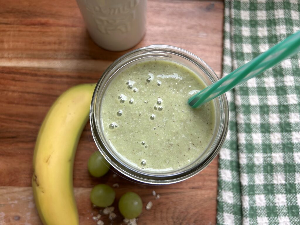 A single creamy green smoothie with oats is served in a clear glass mason jar. Bits of blended seeds are visible in the sage green-colored drink. The smoothie is served on a wooden cutting board with a whole yellow banana, 3 whole green grapes and few scattered whole oats next to the glass for a decorative glance at the ingredients. A green and white checked tea towel is next to the smoothie. 