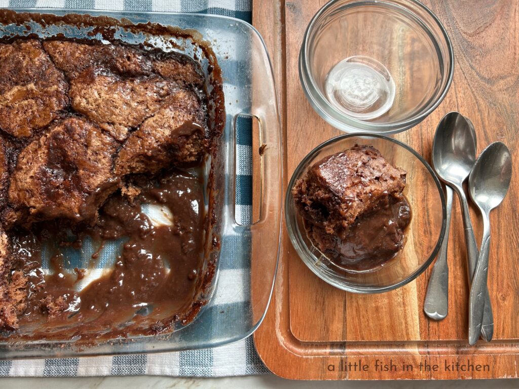 A single of serving of decadent chocolate cobbler is plated in a small, clear glass dessert cup. The crumb on the sides of the homemade chocolate cake look fluffy and moist it's nestled into a  rich puddle of hot chocolate sauce. The glass desserts cups are on a wooden cutting board with 3 spoons just beside the stack of bowls. The clear glass baking dish, containing the whole chocolate, cobbler is just beside the cutting board. A tempting pool of warm chocolate sauce is pooled in the bottom of the baking dish. 