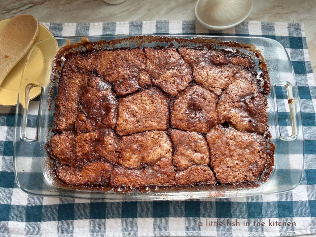 A fresh baked chocolate pudding cake in a clear glass baking dish cools on a rack. The rack is on top of a a blue and white checkered tea towel with a small yellow plate and a wooden spoon laid out beside the baking dish. 