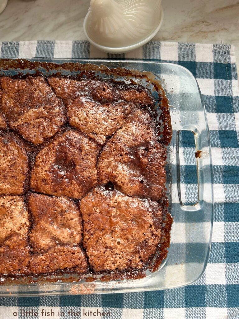 The crackled top of a fresh baked chocolate cobbler, also known as an old-fashioned chocolate pudding cake. Hot fudge sauce is pooled all around the cake in the clear glass baking dish and craters of hot chocolate sauce are pooled in the nooks and crannies of the brownie-like chocolate cake. 