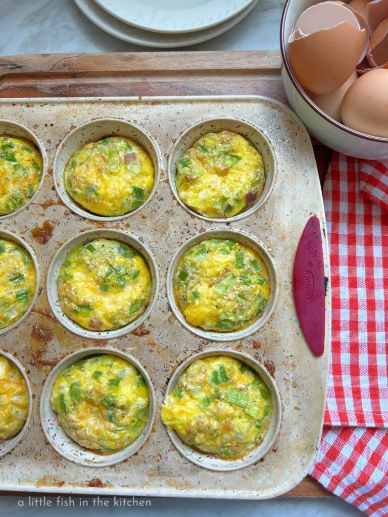 Freshly baked Denver egg muffins filled with ham, diced bell peppers, onions, and melted cheddar cheese cooling in a muffin tin set on a wire cooling rack. The golden egg muffins have lightly browned tops with colorful vegetables visible throughout. There is a red and white checkered kitchen towl just beside the muffin tin and a ceramic bowl filled with a dozen cracked egg shells is next the the warm muffin tin for a decorative effect. 