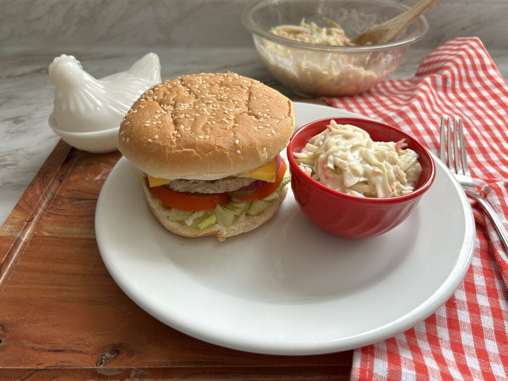 A hearty cheeseburger and a small red ramekin with a serving of prepared, simple creamy coleslaw are served on a white plate. A fork rests on a red and white checkered kitchen towel that is laid out on a wooden cutting board. A clear glass bowl with the prepared batch of slaw and a wooden serving spoon is slightly blurred in the background
