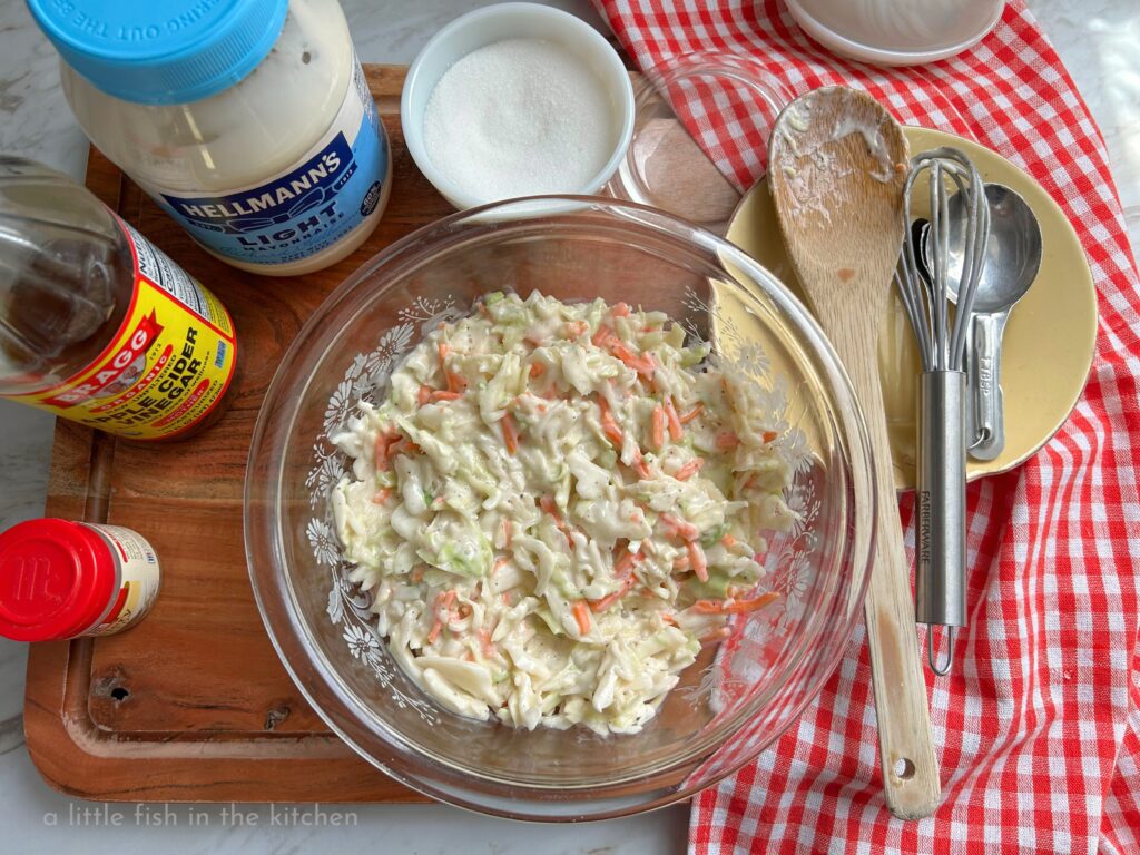 A clear glass bowl is filled with a freshly prepared batch of creamy Southern coleslaw. Shredded cabbage and carrots are generously coated with white creamy dressing. The bowl of slaw is on a large wooden cutting board and surrounded by the gathered recipe ingredients. A jar of mayo, a bottle of vinegar, a small spice jar of celery seed and a small vintage sugar bowl  are on the right of the bowl. On the left a red and white checkered tea towel has a small yellow plate on it with a wooden spoon, a small whisk and measuring spoons resting on it. 