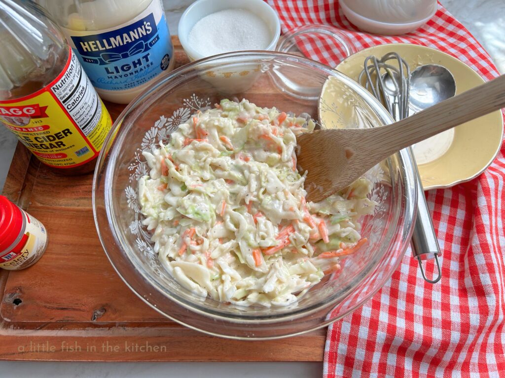 A clear glass bowl is filled with a freshly prepared batch of creamy Southern coleslaw. Shredded cabbage and carrots are generously coated with white creamy dressing. The bowl of slaw is on a large wooden cutting board and surrounded by the gathered recipe ingredients. A jar of mayo, a bottle of vinegar, a small spice jar of celery seed and a small vintage sugar bowl  are on the right of the bowl. On the left a red and white checkered tea towel has a small yellow plate on it with a wooden spoon, a small whisk and measuring spoons resting on it. 