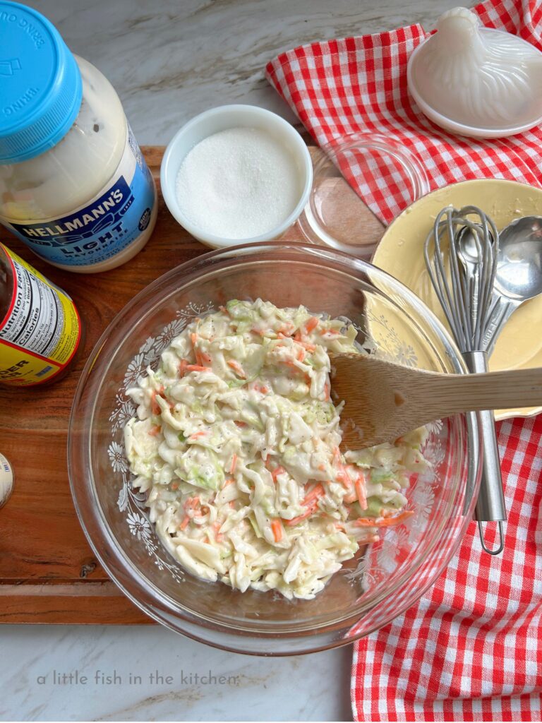 A clear glass bowl is filled with a freshly prepared batch of creamy Southern coleslaw. Shredded cabbage and carrots are generously coated with white creamy dressing. The bowl of slaw is on a large wooden cutting board and surrounded by the gathered recipe ingredients. A jar of mayo, a bottle of vinegar, a small spice jar of celery seed and a small vintage sugar bowl  are on the right of the bowl. On the left a red and white checkered tea towel has a small yellow plate on it with a wooden spoon, a small whisk and measuring spoons resting on it. 