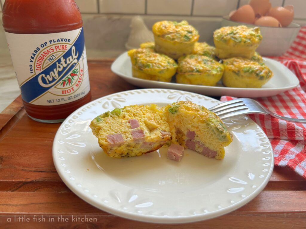 A single Denver egg omelet is on a small white ceramic plate. The egg muffin in cut in half exposing a fluffy egg muffin with melted cheese, bits of smoky ham, green onion and bell pepper visible throughout the savory western-style egg muffin. A red and white checkered tea towel is just beside the platter and plate with egg muffins, and a bottle of Crystal Louisiana hot sauce is next to the plates for serving.