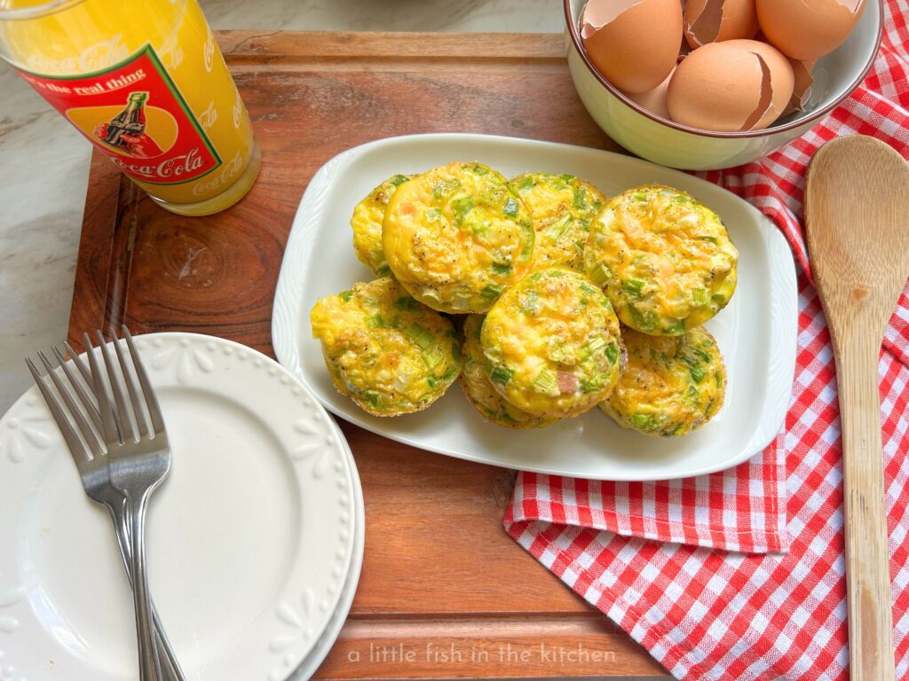 Denver omelette egg muffins filled with diced ham, red and green bell peppers, onions, and melted cheddar cheese, arranged on a rectangular white plate. The fluffy, protein-packed egg muffins have lightly browned tops with colorful vegetables peeking through, making for a hearty and inviting make-ahead breakfast. There is a red and white checkered kitchen towl just beside the muffin tin and a ceramic bowl filled with a dozen cracked egg shells is next the the warm muffin tin for a decorative effect. 