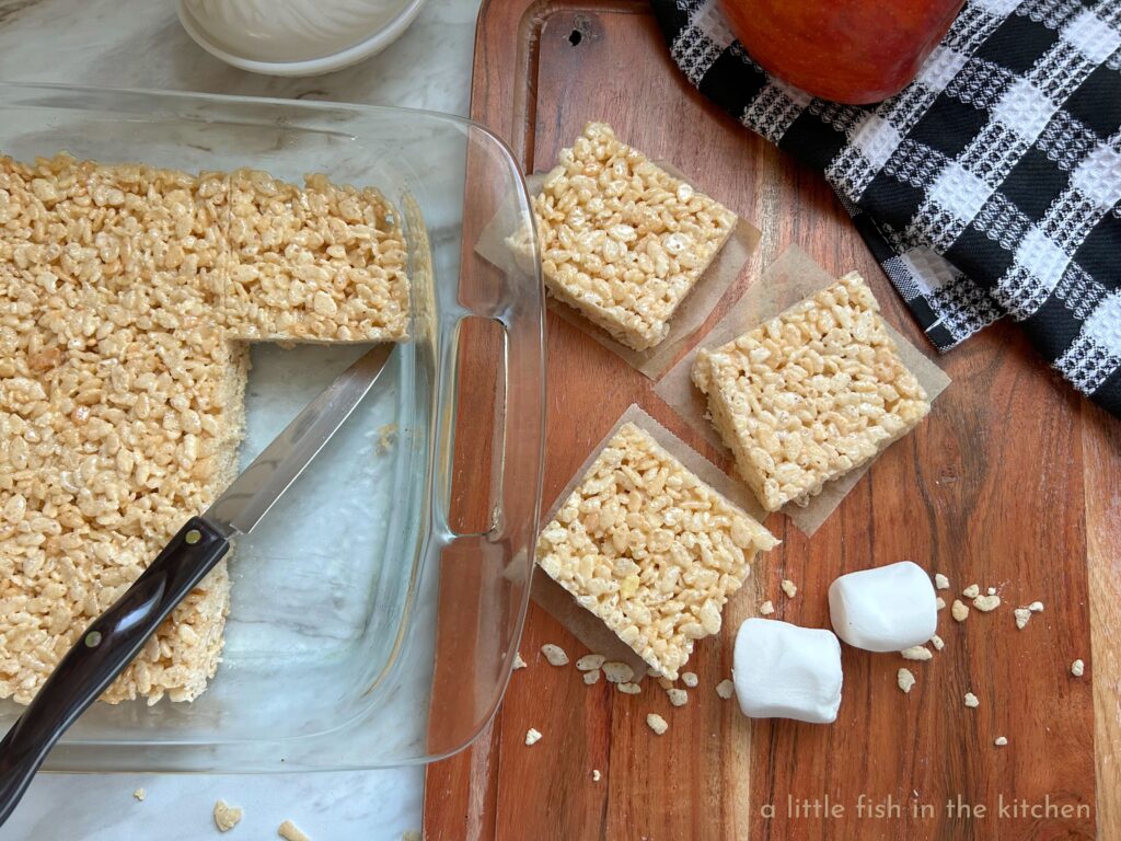 A freshly made batch of classic rice krispie treats is cooled and being cut into squares for serving. The rice krispie treats are in a clear glass baking dish right next to a wooded cutting board. The individual rice krispie treat squares are served on pieces of unbleaches parchment paper. A black and white tea towel rests on the cutting board with a red apple resting on top of the towel. 