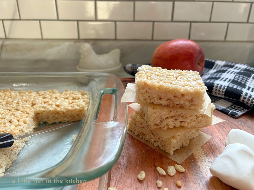 3 freshly prepared classic crispy rice squares are stacked on a wooden cutting board with pieces of unbleached parchment paper between them. 