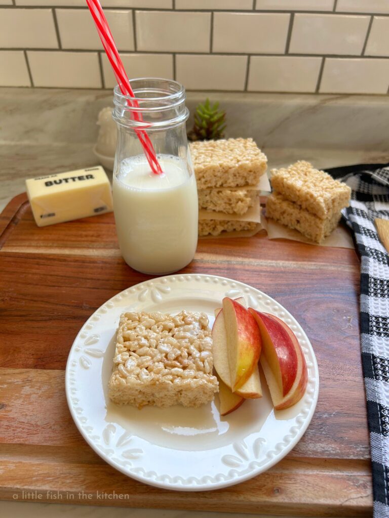  A gooey rice krispie treat is served on a small white ceramic plate with small stack of fresh apple slices next to it for the ultimate after school snack. A clear glass mason jar is half filled with ice-cold milk to enjoy, right next to the plate.