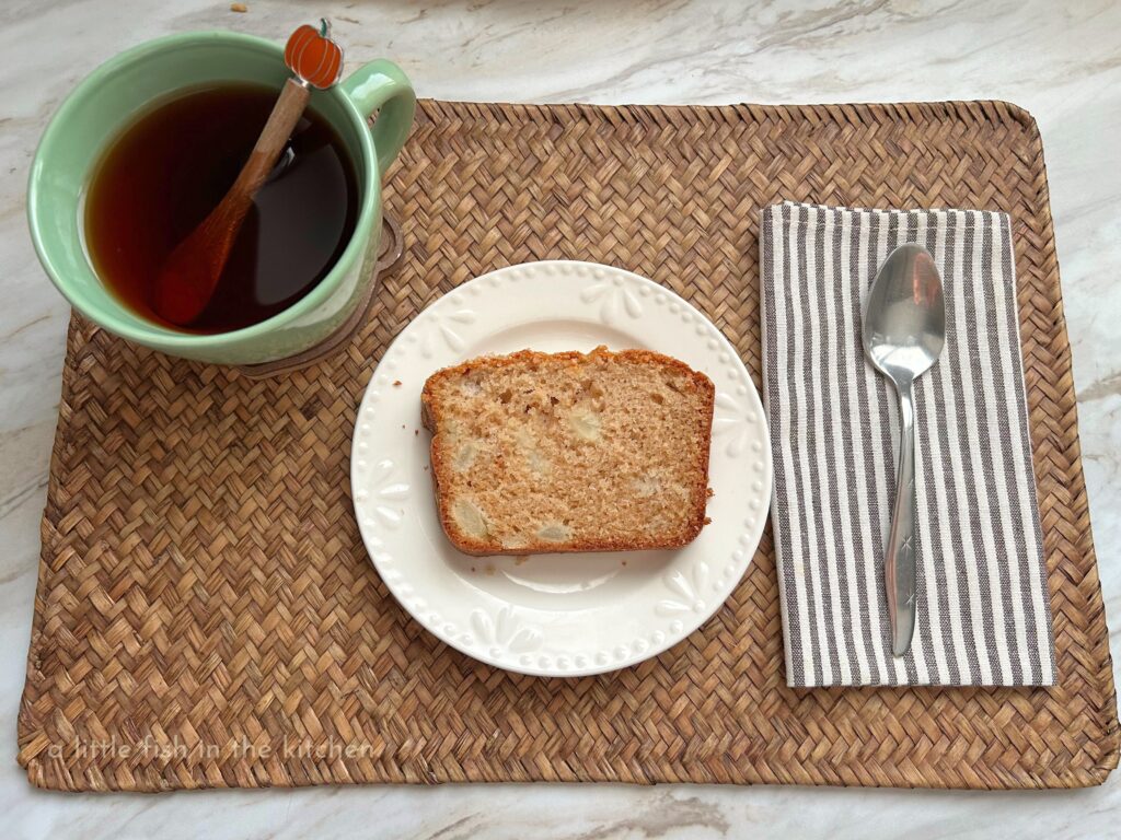 A white dessert plate with a fresh slice of golden, sweet pear quick bread is served on a woven bamboo placemat. Bits of delicious, diced pear are visible within the crumb of the bread. A brown and white striped cloth napkin is neatly folded and topped with a spoon. A green tea cup with freshly steeped English breakfast tea is next two the plate.  