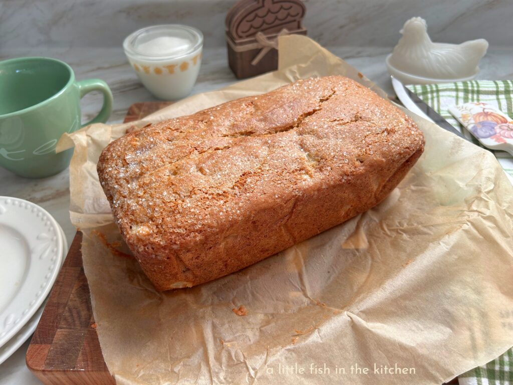 Fresh baked pear quick bread is on a cutting board and still on top of the crinkled parchment paper it was baked in. The top of the pear bread is invitingly crackled and coated with sparkling sugar
