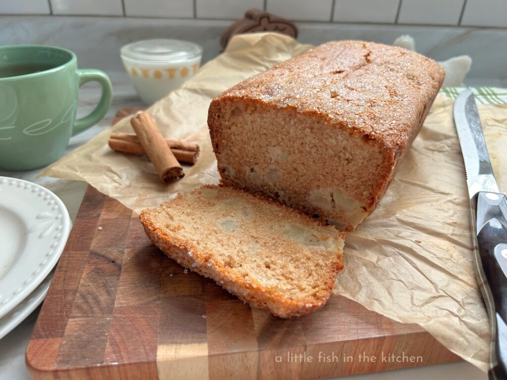 A loaf of spiced pear quick bread is sliced revealing a tender crumb that's studded with sweet bits of pears dotted throughout. The bread is on a wooden cutting with a serrated bread knife next to it. A green tea cup with a steeping tea bag is next to the cutting board along with two white dessert plates.