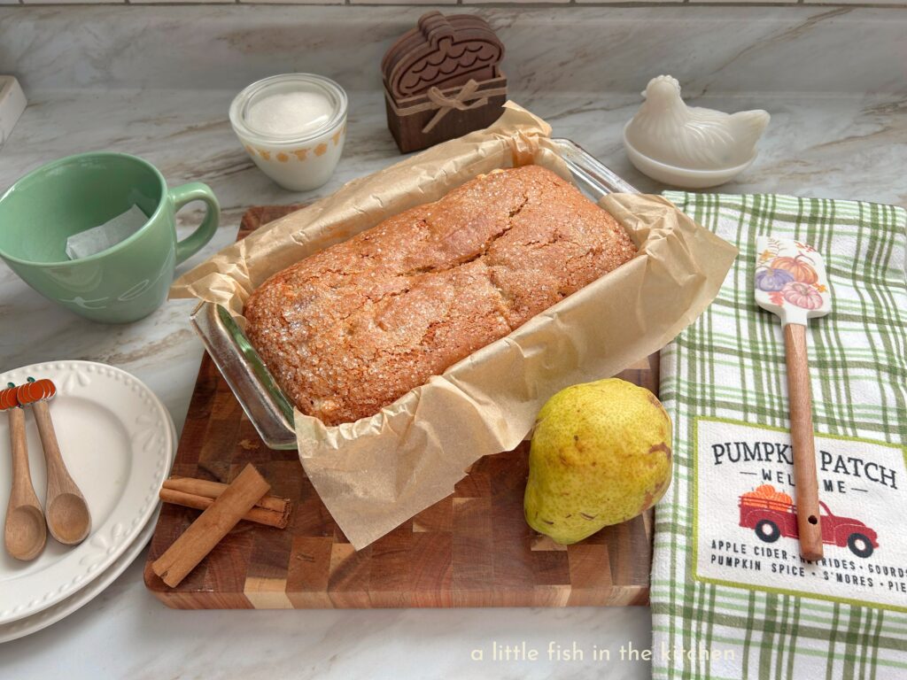 A freshly baked loaf of spiced pear vanilla quick bread rests on a wooden cutting board. The bread is in a clear glass bread dish that's lined with unbleached parchment paper. The top is delightfully crackled and lightly coated with coarse sparkling sugar. There is a whole pears and two whole cinnamon sticks on the cutting board as well for a decorative effect. Beside the cutting board there is a green and white plaid novelty tea towel with a print of a red truck with pumpkins in the back near the bottom of the towel. The words " Pumpkin Patch - Welcome" are printed just above the truck. One the left side of the cutting board there is a light green tea cup with a tea bag inside waiting to be filled with hot water. There are two white ceramic dessert plates with two tiny wooden dematisse spoons with decorative pumpkin topping lying on the plates. 