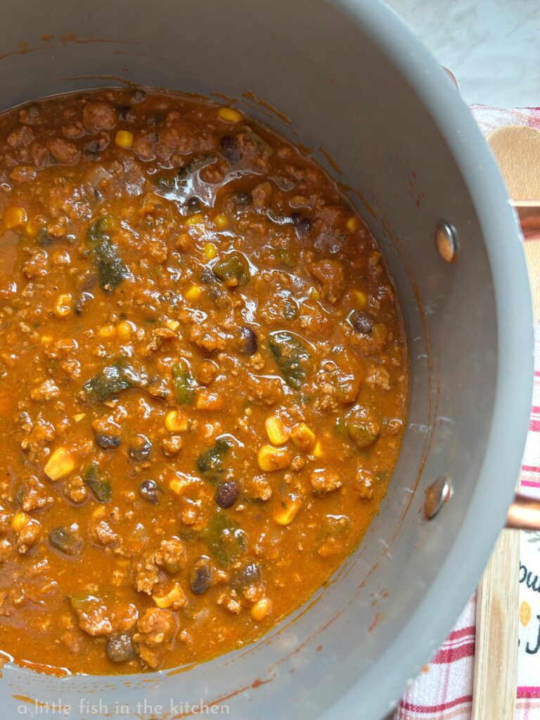 A fresh cooked pot of pumpkin chili with poblano peppers, beans and corn is ready to serve. The amber colored chili looks rich and hearty with bits of ground turkey, green peppers, corn kernels and earthy black beans visible in the mix. 