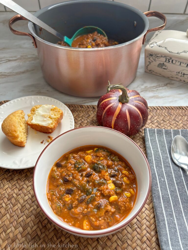 A bowl of flavorful pumpkin chili with poblano peppers is in a white soup bowl with  brown rim. The bowl is on a woven bamboo placemat and a small white appetizer plate with a buttered corn muffins is next to the bowl. A maroon decorative pumpkin is on the placemat in front of a dark gray cloth napkin with a spoon resting on top. The Dutch oven with the rest of the chili is just behind the bowl of chili.