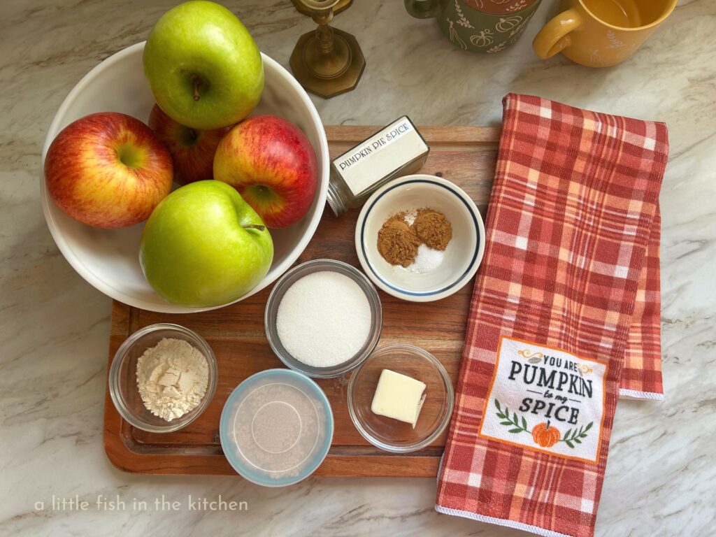 The ingredients are gathered to make a batch of crock pot country apples. A white bowl with six apples is surrounded by ramekins containing spices, white sugar, brown sugar, butter and flour. The ingredients are gathered on top of a rustic wooden cutting board and a decorative orange and white plaid tea towel with the words "you are the pumpkin to my spice" printed on it. 