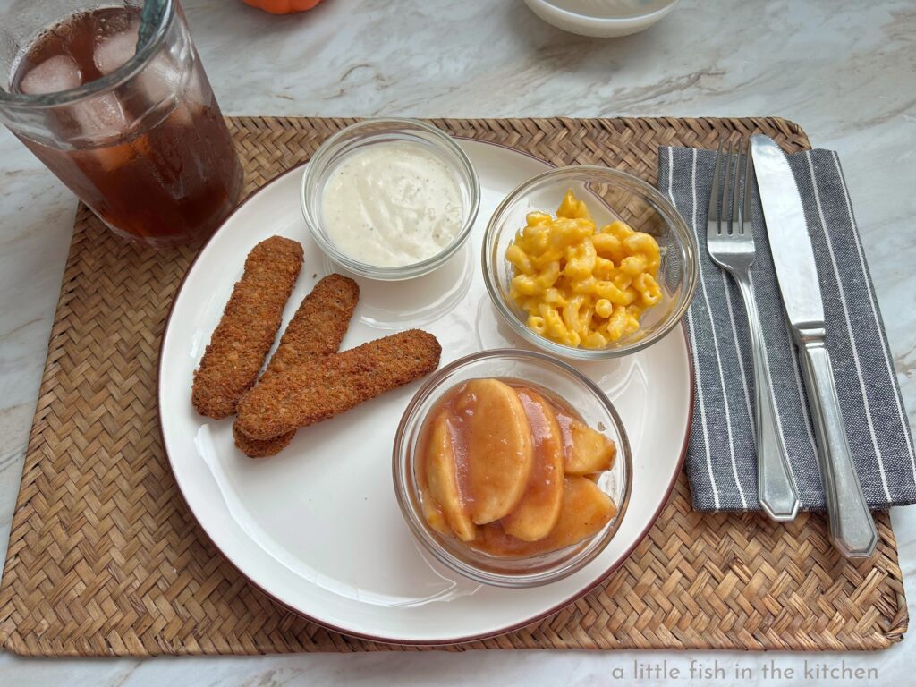 A dinner plate with 3 crispy steak fingers, a small ramekin of mac and cheese, and another ramekin with homemade country apples are served with a chilled glass of iced tea. 