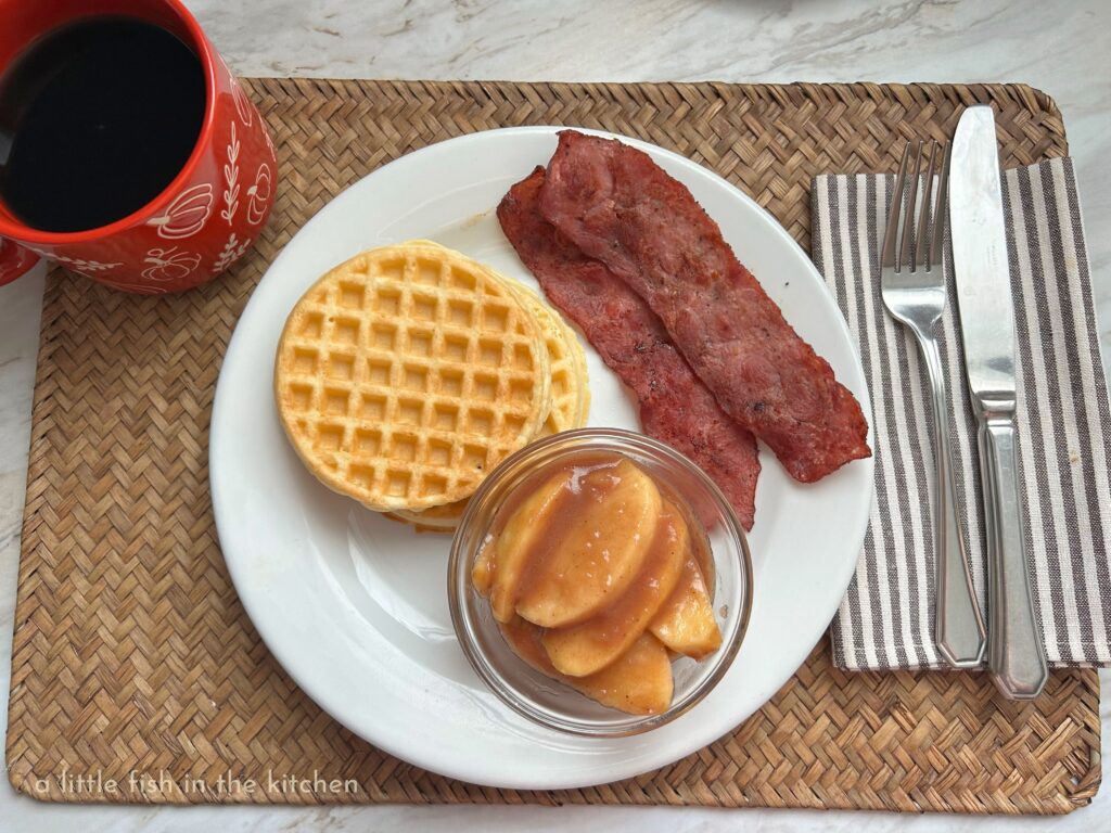Two toasted waffles, two slices of bacon, and a ramekin filled with warm, tender pumpkin spice country apples are served on a white plate for a fantastic breakfast or brunch!