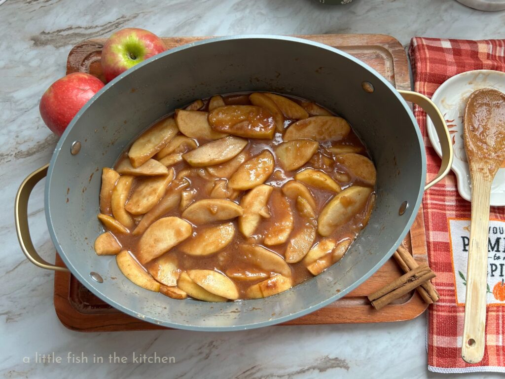An oval-shaped slow cooker crock half-filled with a freshmade batch of pumpkin spice country apples is on a cooling rack. The cooling rack is on top of a wooden cutting board and an orange and white plaid decorative kitchen towel is next to the pot. 