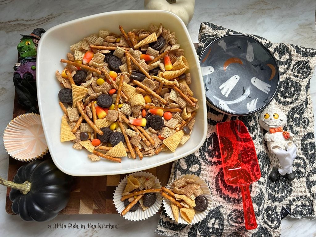 A vintage square serving bowl is filled with an eye-catching batch of no-bake Midnight Moon Munchie Mix. In the bowl, corn chips, candy corn, pretzel sticks, mini Oreo cookies, and candy pieces are all visible. 