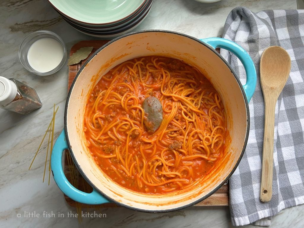 A light blue cast iron Dutch oven is filled with creamy homemade spaghetti. Bits of ground meat are visible mixed in with the tender spaghetti noodles and rich red sauce. A gray and white checkered towel is folded next to the blue Dutch oven with a wooden spoon laying on top. On the other side of the pot a few strands of uncooked spaghetti, a small ramken with cream and a jar  of crushed red pepper flakes are placed for serving and for a deocorative effect. 