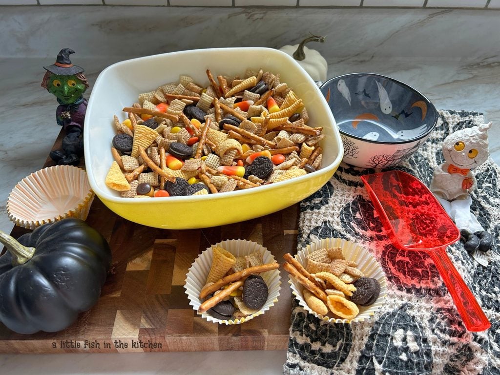 A vintage square serving bowl is filled with an eye-catching batch of no-bake halloween snack mix. In the bowl, corn chips, candy corn, pretzel sticks, mini Oreo cookies, and Reese pieces candy pieces are all visible. 