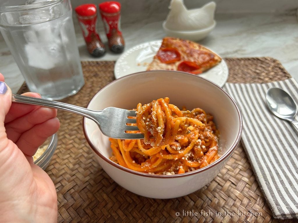 A hand holds a fork with a generous twirl of creamy spaghetti on the end, ready to be enjoyed. Bits of grated parm, ground meat and creamy red sauce are all visible in the pasta dish. A novelty ceramic set of salt and pepper shakers shaped like red and brown cowboy boots are slightly blurred in the photo background. They are above the bamboo placemat. 
