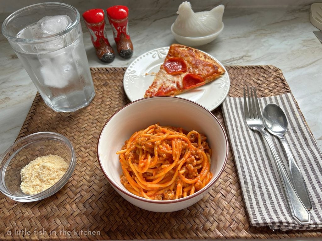 A woven bamboo placemat is set with a white bowl with a brown rim and it in a generous serving of one-pot spaghetti is ready to be enjoyed. A small salad plate with a single piece of pepperoni pizza is also on the placemat next to the bowl of pasta. On the right of the bowl, a brown and white striped cloth napkin is set with a fork and a spoon. on the other side of the bowl, a chilled glass of water and small ramekin with grated parmesan cheese is there ready to serve. 